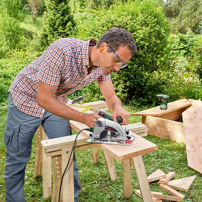Bosch Sierra circular de mano PKS 40 - 850 W, Diámetro hoja de sierra: 130 mm, Profundidad de corte a 90 °: 0 mm - 40 mm - Hombre serrando madera con una sierra circular Bosch en el jardín, con gafas de seguridad y protección auditiva.