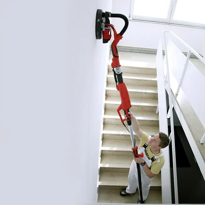Berg Tecmix Wand- & Deckenschleifer LHS 600 - 600 W, Durchmesser Schleifteller: 225 mm - Ein Mann benutzt einen Langhalsschleifer, um eine Wand in einem Treppenhaus zu bearbeiten. Das Werkzeug ist rot und schwarz.