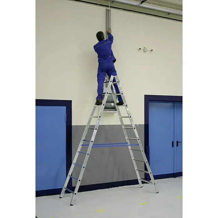 Krause Stabilo Doppelleiter - Anzahl Tritte: 20 Stk., Arbeitshöhe: 4,1 m, Aluminium - Eine Person auf einer KRAUSE Stehleiter, die an einer grauen Wand arbeitet, trägt blaue Arbeitskleidung.