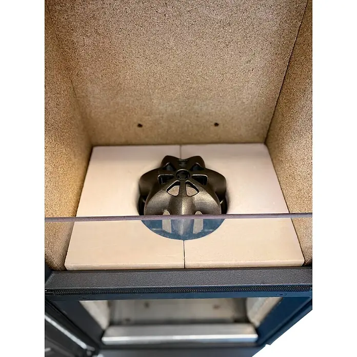 xeoos Kaminofen Patagonia X5 - 5 kW, Raumheizvermögen: 20 m³ - 70 m³, Farbe Verkleidung: Inox Edelstahl - Innenansicht eines Kaminofens mit einem stilvollen, dunkelgrauen Einsatz und hellen, quadratischen Keramikplatten als Basis. Die Konstruktion wirkt robust und modern.