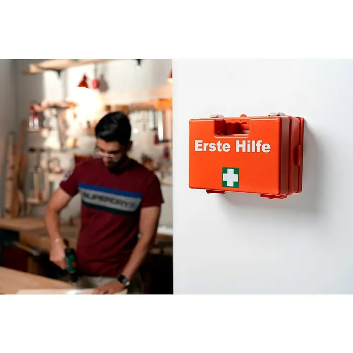 Leina-Werke Erste-Hilfe-Koffer Quick - DIN 13157, Mit Wandhalterung, Orange - Erste Hilfe Kasten in leuchtendem Orange mit deutlichem grünen Kreuz und weißer Beschriftung "Erste Hilfe" an einer hellen Wand montiert.