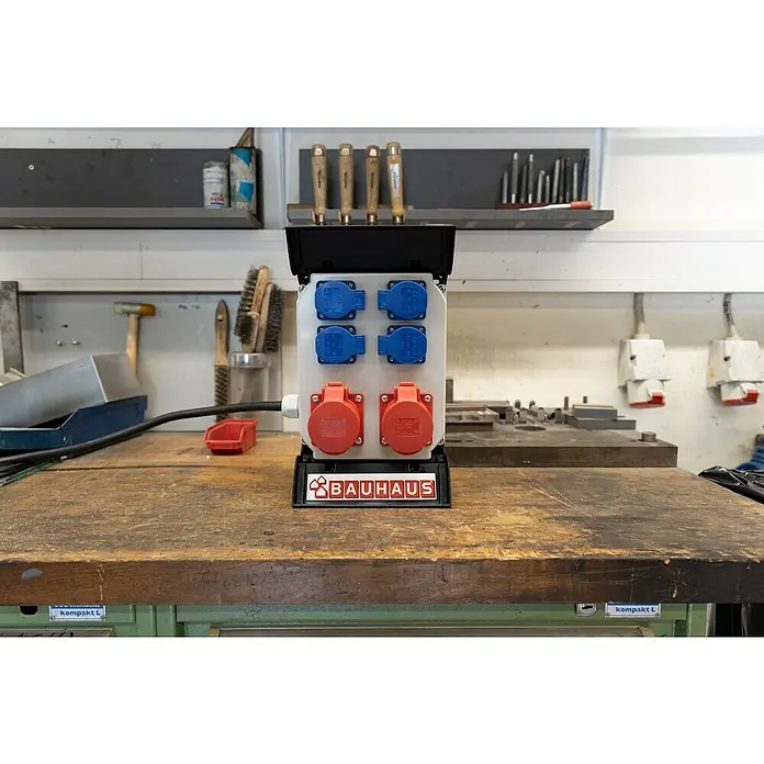 BAUHAUS Baustromverteiler - Anzahl CEE-Steckdosen: 2 Stk., Anzahl Steckdosen Schuko: 4 Stk., IP44 - Eine Mehrfachsteckdose mit roter und blauer Farbgebung auf einer Werkbank, versehen mit einem "Bauhaus"-Logo für den Heimwerkerbedarf.