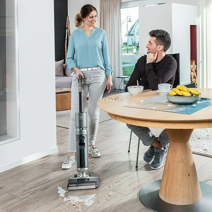 Thomas Akku-Hartbodenreiniger Aqua Floor Cleaner Cordless - 21,6 V - Eine Frau benutzt einen kabellosen Staubsauger, um eine Flüssigkeit auf einem Holzboden aufzusaugen, während ein Mann zusieht.