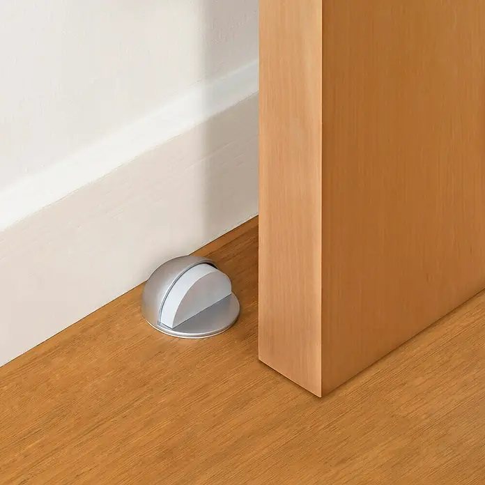 Inofix Tope de puerta 2099 - Blanco - Primer plano de un tope de puerta plateado en un suelo de madera contra una puerta y un zócalo blanco.