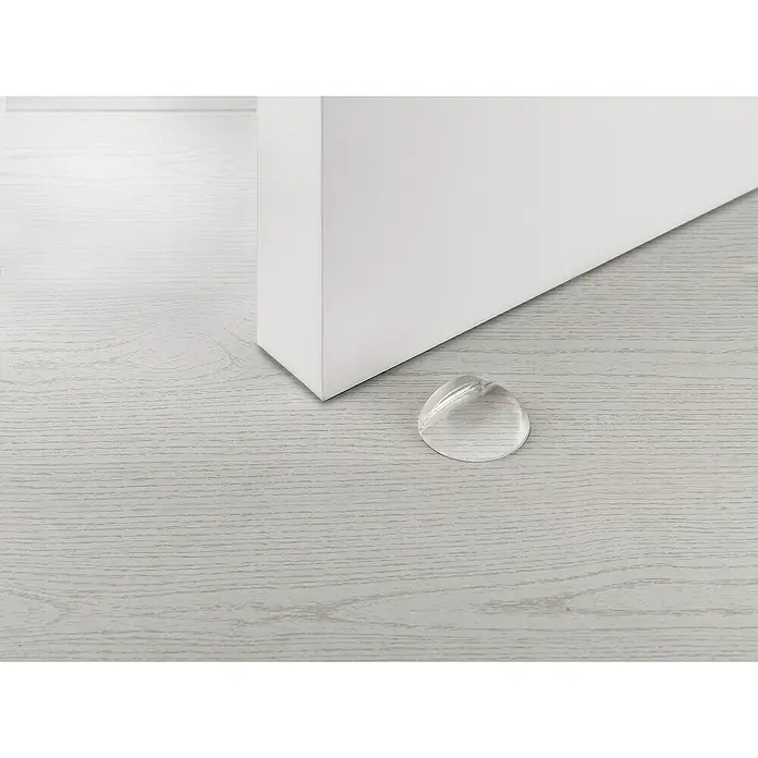 Inofix Tope de puerta 2050 - Caucho macizo, Transparente, 4 ud. - Esta imagen muestra un tope de puerta transparente colocado en un suelo de madera clara, protegiendo una puerta blanca de daños.