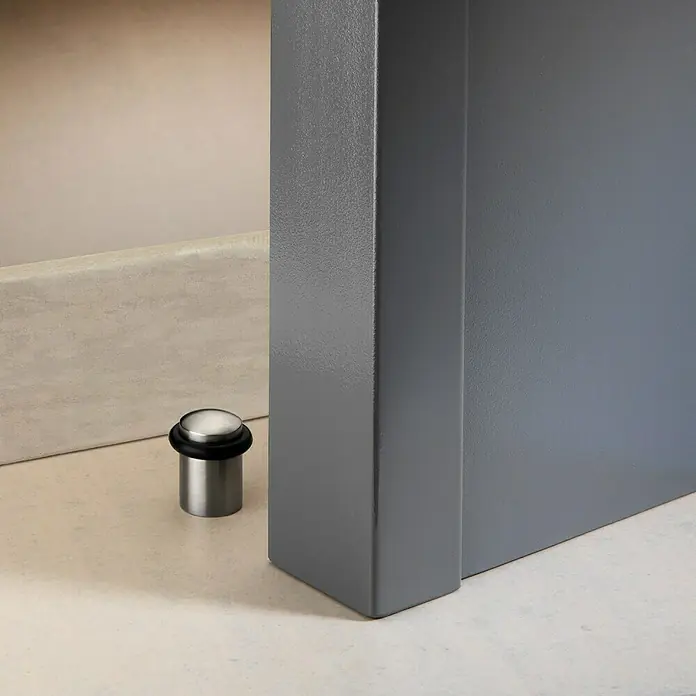 Inofix Tope de puerta 3160 con tórica - Acero inoxidable, Plateado/Negro, 6 ud. - Primer plano de un tope de puerta de acero inoxidable con un anillo de goma negra, sobre un marco de puerta gris.