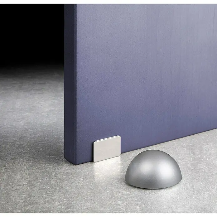 Inofix Tope de puerta 2034 - Plástico, Cromo, 1 ud. - Detalle de un tope de puerta plateado y un elemento de fijación contra una pared morada. El tope de puerta tiene forma de cúpula.