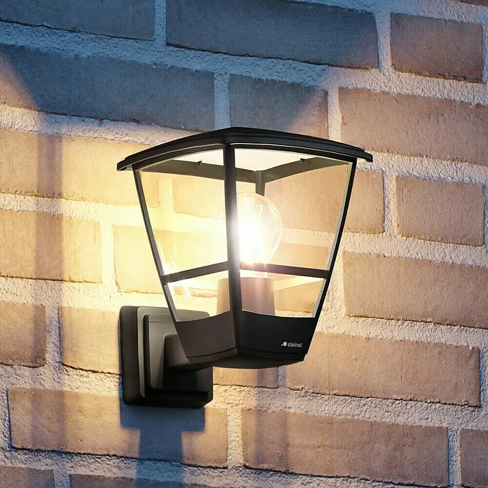 Steinel Außenwandleuchte L 10 - 15 W, 22,4 x 17,5 x 24,2 cm, Schwarz, IP44 - Eine moderne, schwarze Steinel Außenwandleuchte mit klarem Design und warmem Licht, montiert an einer Ziegelwand.