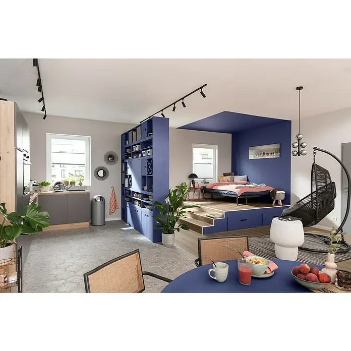 SCHÖNER WOHNEN-Farbe Wandfarbe Designfarbe - Sanftes Seidengrau, Matt, 2,5 l, Konservierungsmittelfrei - Helle Wohnung mit erhöhtem Schlafbereich, abgetrennt durch ein blaues Regal. Der Essbereich zeigt einen blauen Tisch mit Frühstücksutensilien.