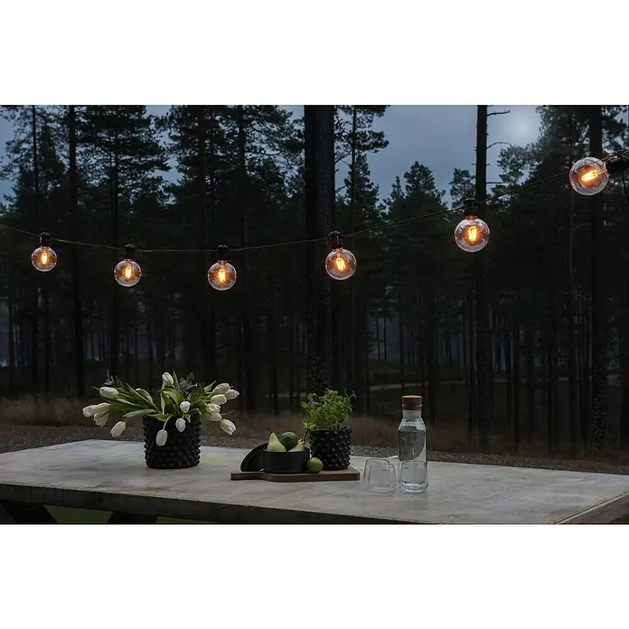 Konstsmide LED-Lichterkette - Außen, 4,5 cm, Bernstein, Rund, Solarbetrieben - Lichterkette mit Edison-Glühbirnen beleuchtet einen Tisch mit Blumen, Pflanzen, Obst, Wasserflasche und Gläsern.