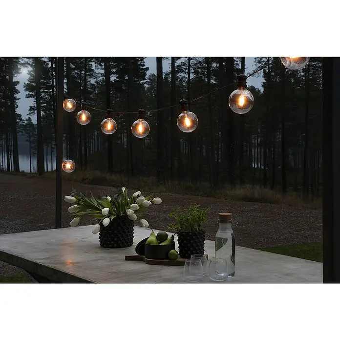Konstsmide LED-Lichterkette - Außen, 4,5 cm, Bernstein, Rund, Solarbetrieben - Eine stimmungsvolle Szene mit dekorativen Glühbirnen, die eine gemütliche Atmosphäre über einem gedeckten Tisch im Freien schaffen.