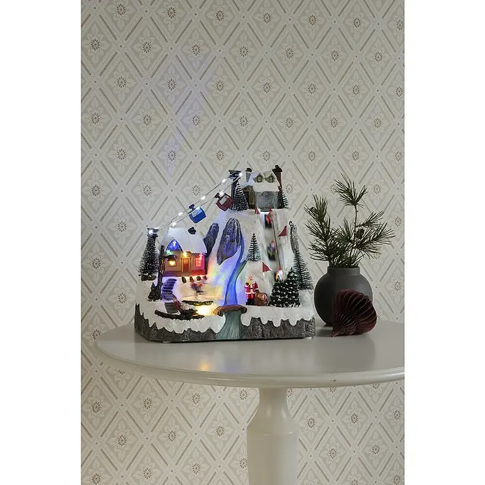 Konstsmide LED-Szenerie Skipiste - L x B x H: 29 x 20,5 x 28 cm, Bunt, Batteriebetrieben, LED - Ein festliches beleuchtetes Weihnachtsdorf-Display auf einem runden Tisch mit einem strukturierten Hintergrund.