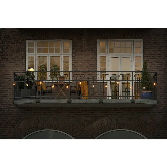 Konstsmide LED-Lichterkette - Außen, 4,5 cm, Bernstein, Oval, Solarbetrieben - Balkon mit Lichterkette, Möbeln und Pflanzen. Die warmen Lichter schaffen eine gemütliche Atmosphäre auf dem Balkon des Backsteingebäudes.