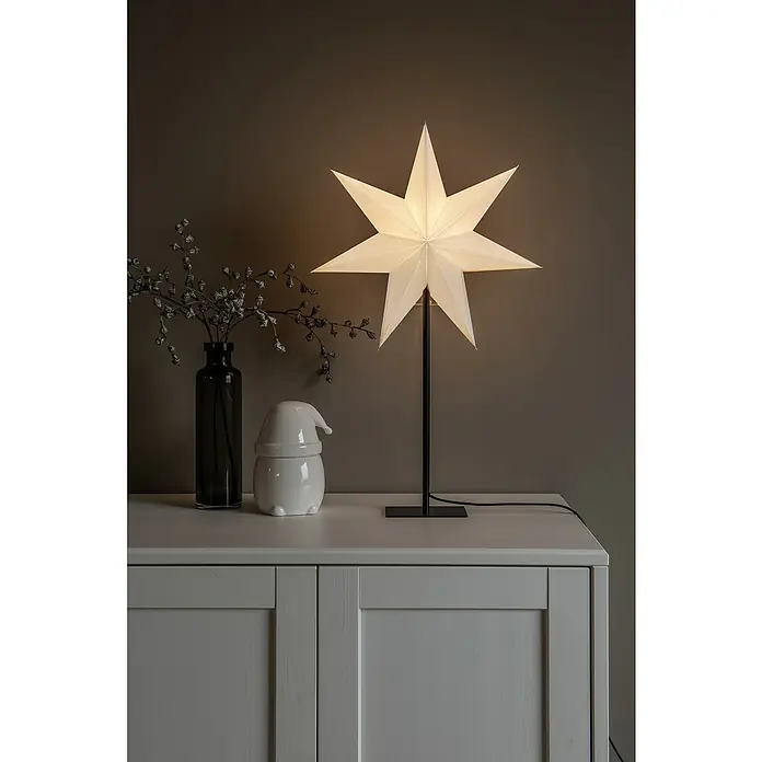Konstsmide Standstern 7 Zacken - Weiß, Schwarz, Netzbetrieben - Sternförmige Lampe auf einer weißen Kommode mit einem kleinen Weihnachtswichtel und einer Vase mit Zweigen.