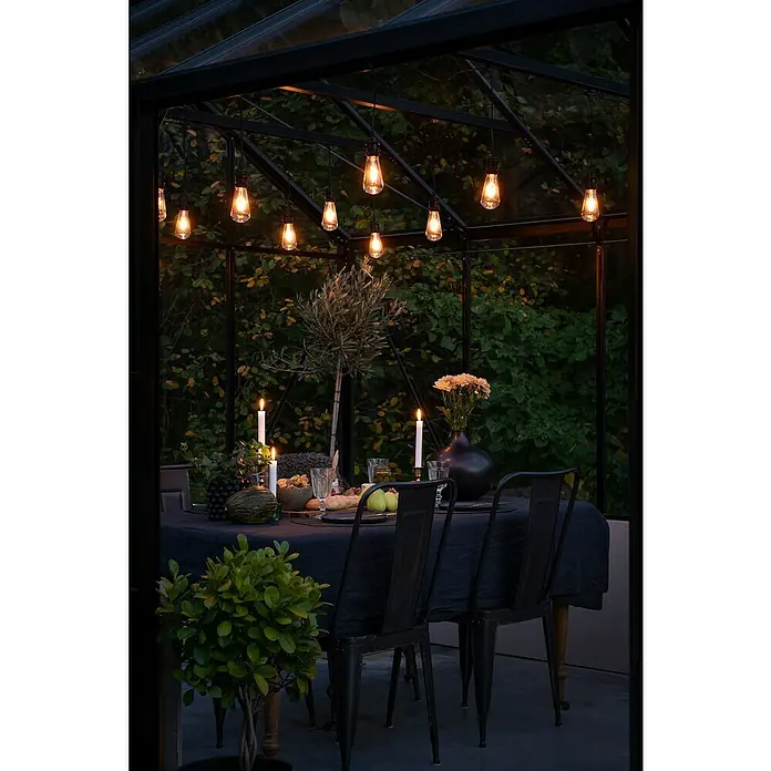 Konstsmide LED-Lichterkette - Länge: 10,85 m - Ein stimmungsvolles Gewächshaus mit einer festlichen Tafel, beleuchtet durch hängende Edison-Glühbirnen, Kerzen und einem elegant gedeckten Tisch.