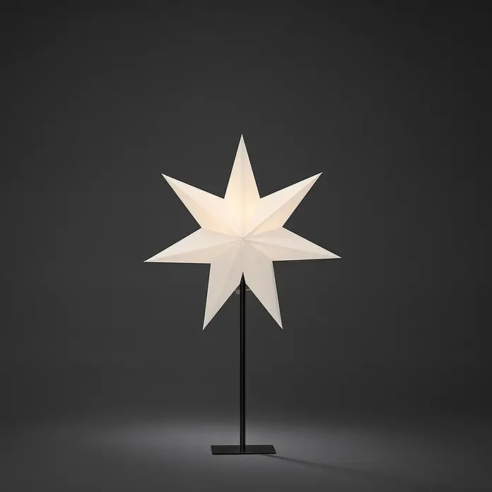 Konstsmide Standstern 7 Zacken - Weiß, Schwarz, Netzbetrieben - Eine elegante Papierstern-Lampe auf einem schlanken, schwarzen Ständer, die warmes Licht in einem dunklen Raum verbreitet.