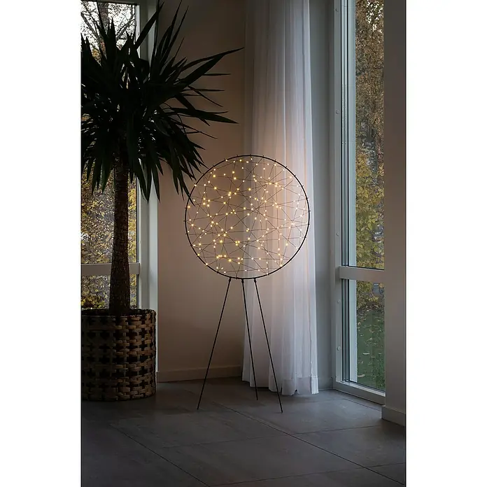 Konstsmide LED-Metallsilhouette Ring - 60 x 25 x 117 cm, Schwarz, Netzbetrieben, Lichtfarbe: Bernstein - Elegante Stehlampe mit kreisförmigem Design und zarten Lichtern, die eine warme Atmosphäre schafft, neben einer Zimmerpflanze.