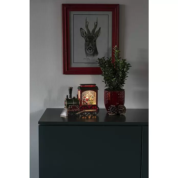 Konstsmide LED-Schneelaterne Zug - Innen, Rot, Grün, 25 x 9 x 19,5 cm, Zug mit Weihnachtsmann, Batteriebetrieben - Eine festliche rot-grüne Weihnachtszug-Dekoration mit beleuchteter Weihnachtsmann-Schneekugel, auf einem dunklen Schrank als Teil einer Weihnachtsdekoration.