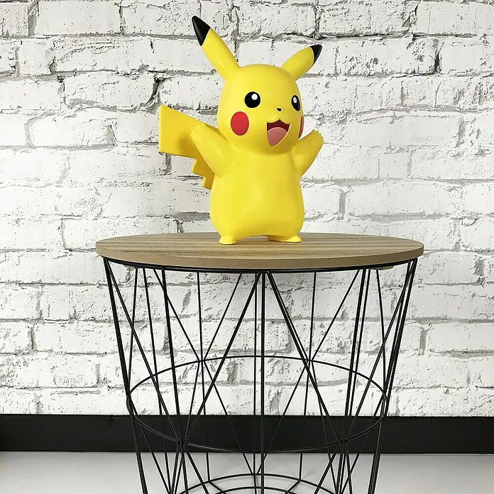 Lámpara LED decorativa Pikachu - L x An x Al: 12,5 x 19 x 25 cm - Una figura amarilla de Pikachu está de pie sobre una mesa auxiliar decorativa con una estructura de alambre geométrica, con una pared de ladrillo blanco.