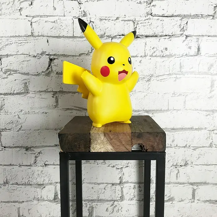 Lámpara LED decorativa Pikachu - L x An x Al: 12,5 x 19 x 25 cm - Figura amarilla de Pikachu con los brazos levantados expuesta sobre una pequeña mesa de madera con estructura metálica negra.