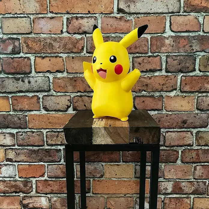 Lámpara LED decorativa Pikachu - L x An x Al: 12,5 x 19 x 25 cm - Figura de Pikachu amarilla con expresión alegre y brazos levantados, sobre un bloque de madera oscuro contra una pared de ladrillo.
