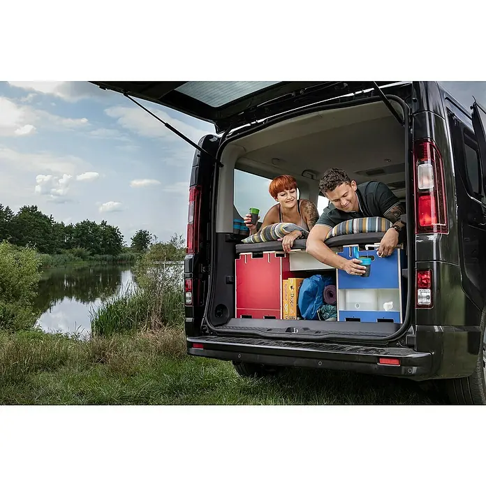Vojo Camper Ausbau-Set Nomad - 23 -tlg., Geeignet für: Transporter - Ein umgebauter Van mit Heckküche und zwei Personen im Innenraum, die die Aussicht auf einen See genießen.