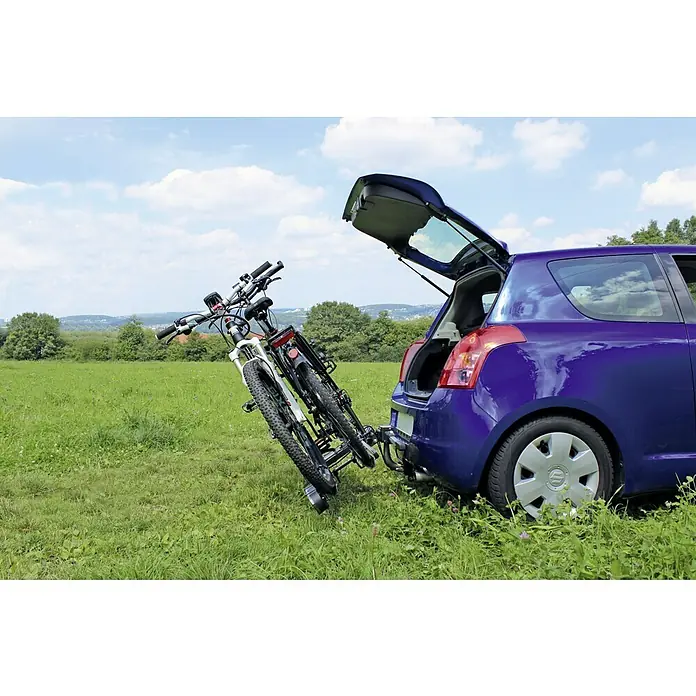 Eufab Portabicicletas Poker-F - Apto para: 2 bicicletas, Capacidad de carga: 60 kg, Específico para: Vehículos con enganche para remolque - Dos bicicletas unidas a un portabicicletas montado en un enganche en un Suzuki Swift azul, con el maletero abierto, sentadas frente a un prado verde y árboles.
