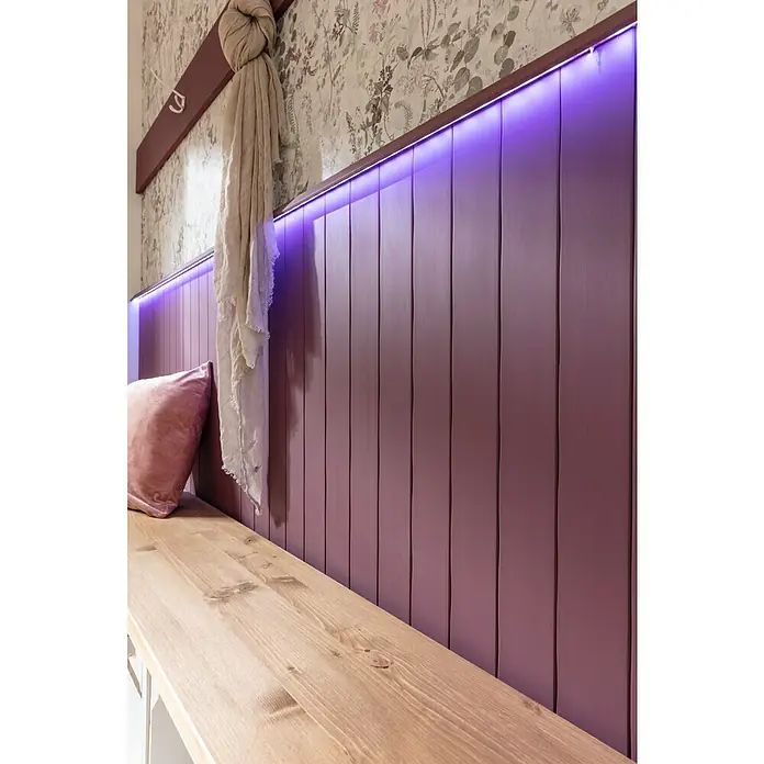 Nmc Decoflair Wandpaneel CP1 - 5 Stk., Weiß, 1,22 m x 79 mm, Hochverdichtetes extrudiertes Polymer (HDPS) - Nahaufnahme eines lila Wandpaneels mit Holzbank und weichem Kissen, beleuchtet mit violettem Licht, das ein gemütliches Ambiente schafft.