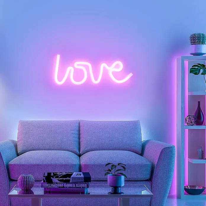 Just Light LED-Dekoleuchte Neon Love - Neon Love, L x B x H: 8,5 x 29,5 x 24 cm, Netzbetrieben - Stilvolles Interieur mit einem grauen Sofa und einem leuchtenden "Love"-Schriftzug an der Wand, akzentuiert durch violette Beleuchtung.