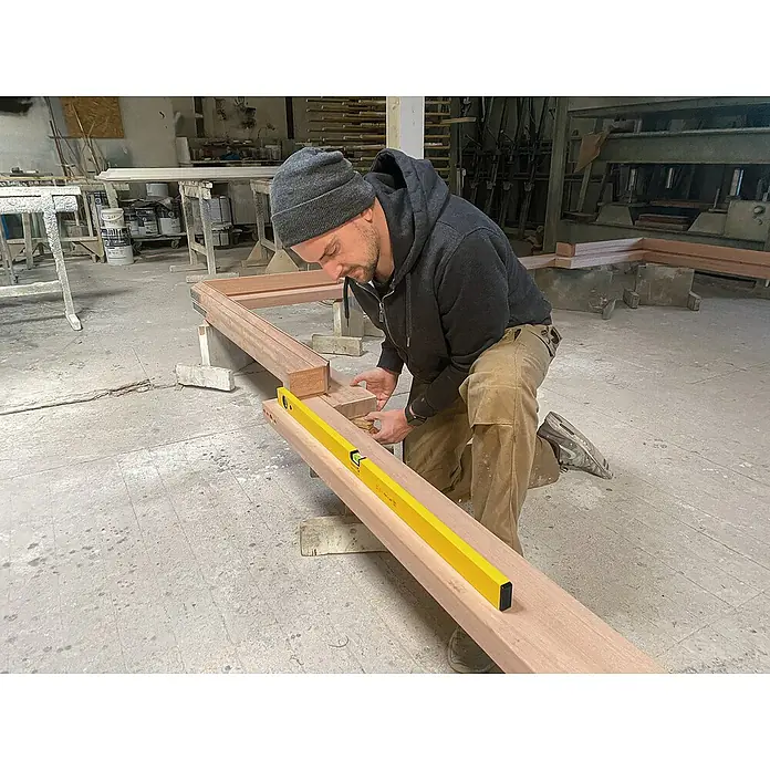 Stabila Wasserwaage Type 70Länge: 90 cm, Aluminium - Ein Handwerker prüft mit einer gelben Wasserwaage die Ebenheit eines Holzrahmens in einer Werkstatt, um präzise Bauarbeiten zu gewährleisten.
