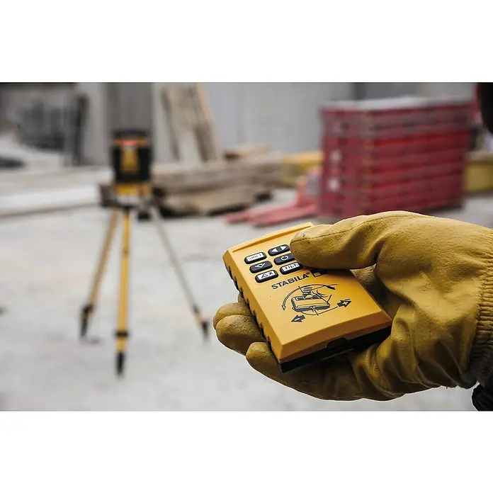 Stabila Rotationslaser LAR 35013 -tlg. - Ein gelber Stabila Laser-Fernbedienung wird von einer behandschuhten Hand gehalten, im Hintergrund ein Laser-Nivelliergerät auf einem Stativ.