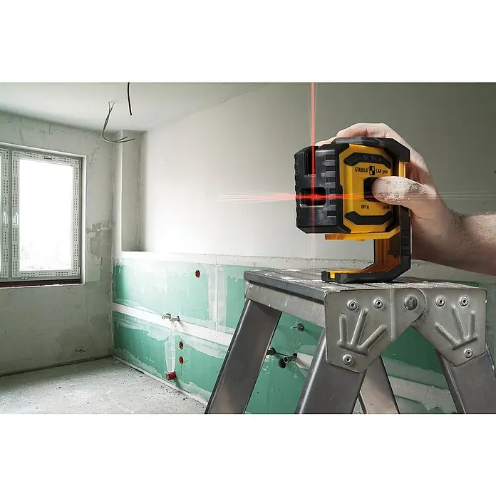 Stabila Kreuzlinienlaser LAX 300 - Max. Arbeitsbereich: 20 m, 8 -tlg. - Baustellenszene mit einem gelben Stabila LAX 300 Kreuzlinienlaser auf einer Leiter, der rote Laserlinien an die Wand projiziert.