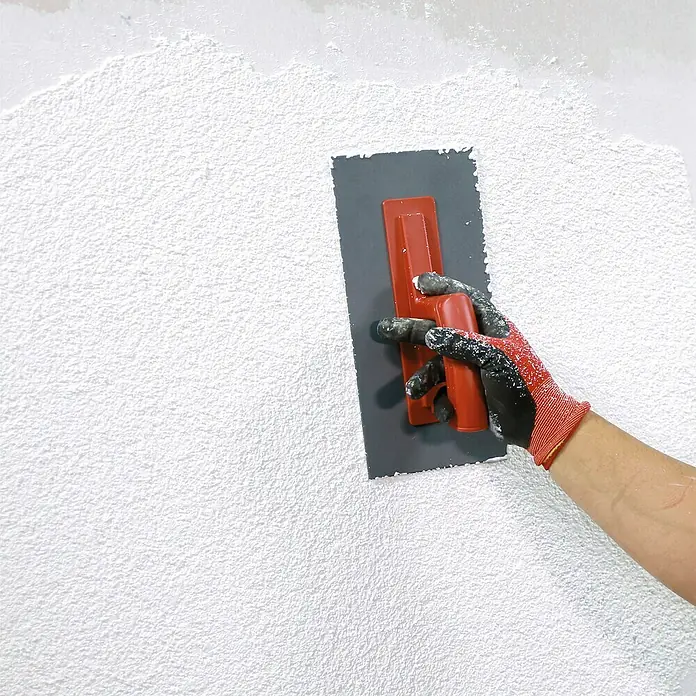 SG Weber Scheibenputz - Körnung: 3 mm, 25 kg - Aufbringen von Strukturputz auf eine Wand mit einer Glättkelle. Die Kelle hat einen grauen Körper und einen roten Griff.