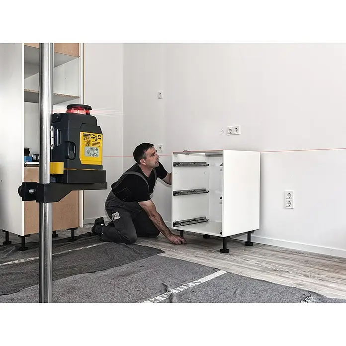 Stabila Kreuzlinienlaser LAX 400 - 10 -tlg., Arbeitsbereich: 20 m - Ein Handwerker nivelliert mit einem Rotationslaser einen weißen Küchenschrank mit schwarzen Füßen und Metallschienen in einem hellen Raum.