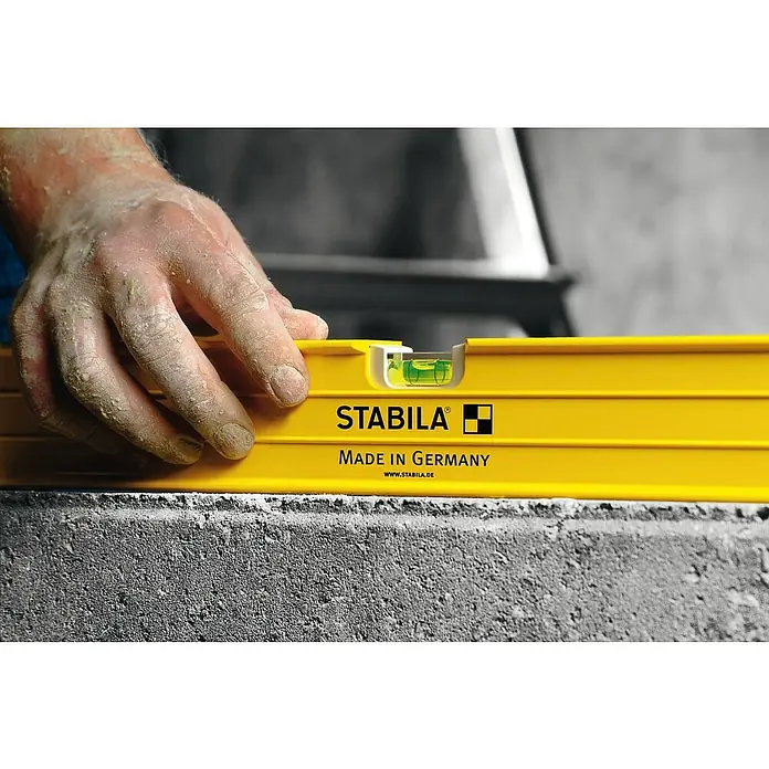 Stabila Wasserwaage Type 96-2Länge: 61 cm, Anzahl Libellen: 3 Stk. - Nahaufnahme einer gelben Stabila Wasserwaage auf einer Betonoberfläche, gehalten von einer Hand, die die Präzision beim Nivellieren betont.