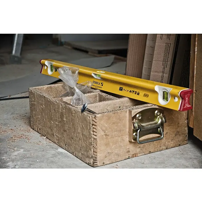 Stabila Wasserwaage Type R 300 - Länge: 244 cm, Anzahl Libellen: 3 Stk. - Eine gelbe Stabila Wasserwaage liegt auf einer Holzkiste, zusammen mit einer Tüte und einem Nagel. Der Kasten dient zur Aufbewahrung.