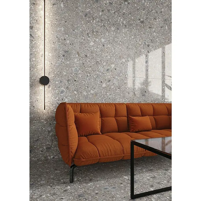 Feinsteinzeugfliese Terrazzo Ceppo - 119,7 x 119,7 cm, Silber, Glänzend - Stilvolles Wohnzimmer mit Terrazzoboden und -wand, ergänzt durch ein modernes orangefarbenes Sofa und eine minimalistische Lampe.