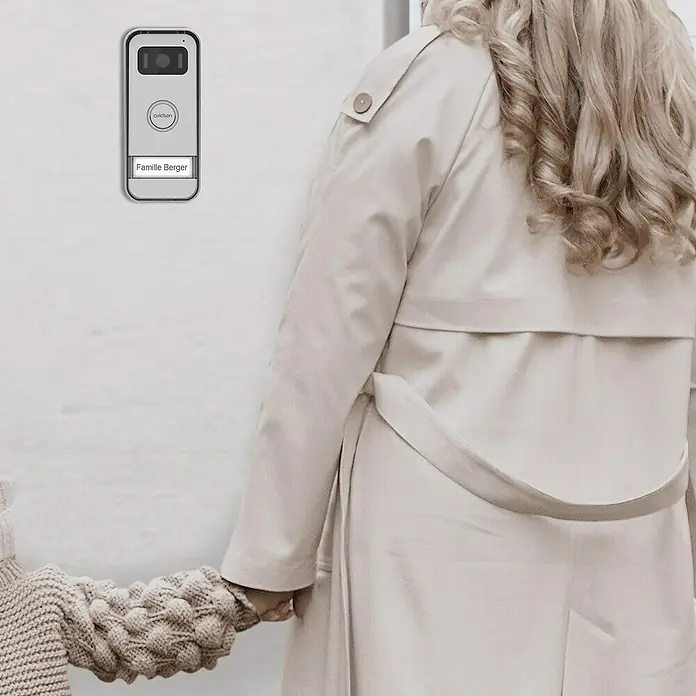 Avidsen Türklingel mit Kamera Video Türsprechanlage MayaSilber, 800 x 480 Pixel - Die Avidsen Smart Türsprechanlage mit Kamera und Namensschild "Familie Berger" ist an einer weißen Wand neben einer Frau montiert.