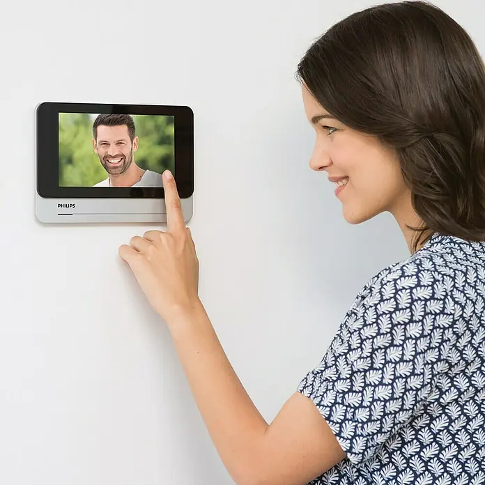 Philips WelcomeEye Farb-Video-Türsprechanlage Wireless - Bildschirmgröße: 7″, 1.920 x 1.080 Pixel (Full HD), Smarte Steuerung: Philips WelcomeEye App - Frau interagiert mit einer Philips Gegensprechanlage. Das Gerät zeigt ein lächelndes Gesicht und ermöglicht eine moderne Hauskommunikation.