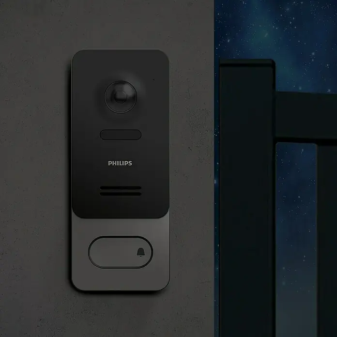 Philips WelcomeEye Farb-Video-Türsprechanlage Wireless - Bildschirmgröße: 7″, 1.920 x 1.080 Pixel (Full HD), Smarte Steuerung: Philips WelcomeEye App - Eine Philips-Video-Türklingel mit Kamera, Mikrofon und Klingelknopf, montiert an einer grauen Wand, neben einem dunklen Zaun bei Nacht.