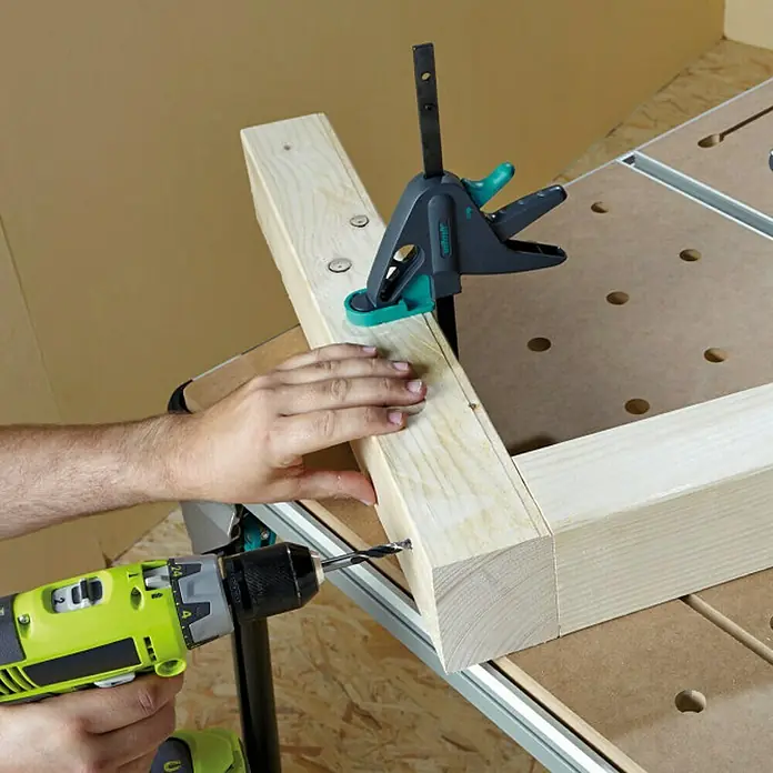 Wolfcraft Sargento 65-150-W - Anchura de sujeción: 120 mm - Artesano usando un taladro inalámbrico para hacer un agujero en un pedazo de madera, que está fijado en un banco de trabajo con una abrazadera.