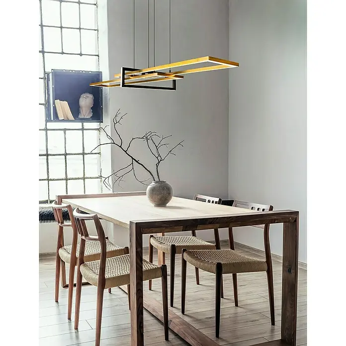Trio Leuchten LED-Pendelleuchte Salinas - 34 W, L x B x H: 40 x 110 x 150 cm, Messing-matt, Warmweiß - Eine rechteckige LED-Pendelleuchte in Gold hängt über einem Esstisch mit Holzstühlen und einem Keramikgefäß mit Zweigen.