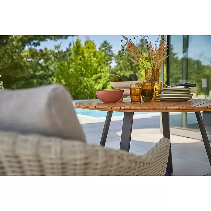 Sunfun Carina Gartentisch rund - Ø x H: 115 x 76 cm, Akazienholz, Teak - Runder Esstisch für draußen mit Holzlattenplatte und dunklen Beinen, gedeckt mit Geschirr und Gläsern auf einer Terrasse am Pool.