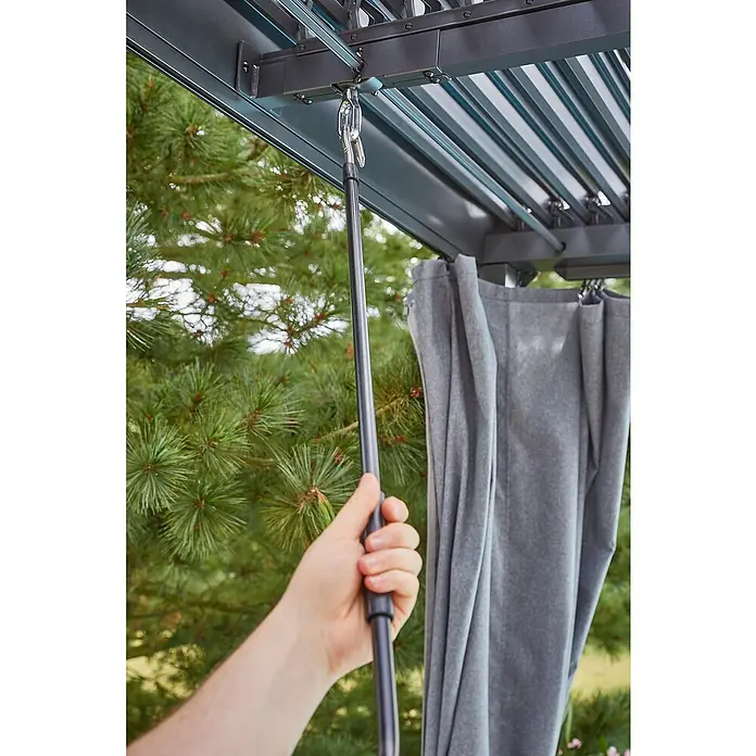 Sunfun Maja Loungeliege - L x B x H: 222 x 159 x 230 cm, Metall, Grau - Nahaufnahme einer Hand, die eine Stange bedient, um die Lamellen eines Outdoor-Pavillons zu verstellen, mit grauem Vorhang im Hintergrund.