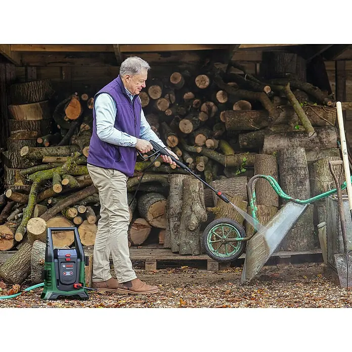 Bosch Universal Akku-Hochdruckreiniger UniversalAquatak 36V-100 - 8 -tlg., 36 V, 1 Akku - Ein Mann reinigt eine Schubkarre mit einem grünen Bosch Hochdruckreiniger vor einem Holzlager.