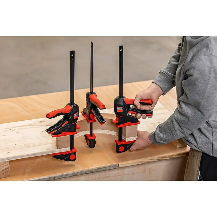 Bessey Einhandzwinge EZ 360-30Spannweite: 300 mm, Ausladung: 82 mm - Drei BESSEY Einhandzwingen auf einer Werkbank, die ein Werkstück fixieren, wobei eine von einer Person bedient wird.