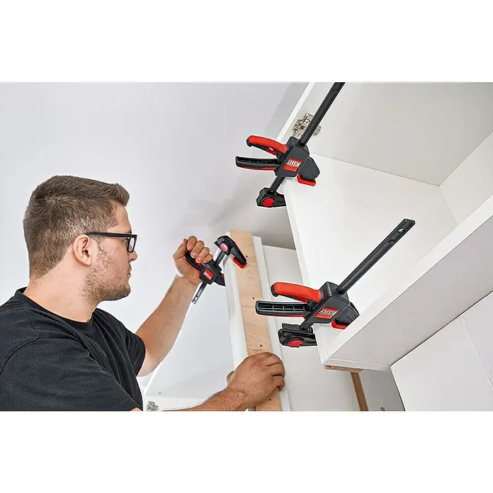 Bessey Einhandzwinge EZR - Spannweite: 150 mm, Ausladung: 60 mm - Der Mann befestigt eine Holzplatte mit BESSEY Einhandzwingen an einem Schrank. Die Zwingen sind rot und schwarz.