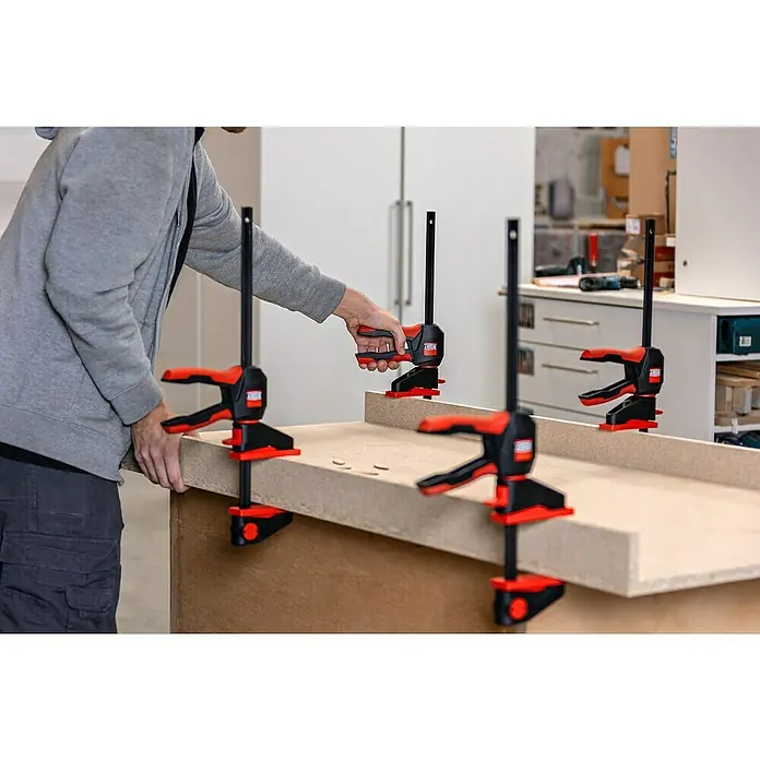 Bessey Einhandzwinge EZ 360-60Spannweite: 600 mm, Ausladung: 80 mm - Ein Handwerker befestigt ein Werkstück mit mehreren rot-schwarzen Einhandzwingen auf einer Werkbank. Die Zwingen fixieren das Material präzise.