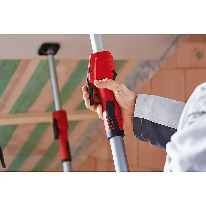 Bessey Montagestütze STE 300Verstellbar: 170 cm - 300 cm - Blick auf eine Hand, die ein rotes BESSEY-Werkzeug zur Deckenstützung hält, während sie eine Decke befestigt, wobei eine zweite Stütze im Hintergrund sichtbar ist.