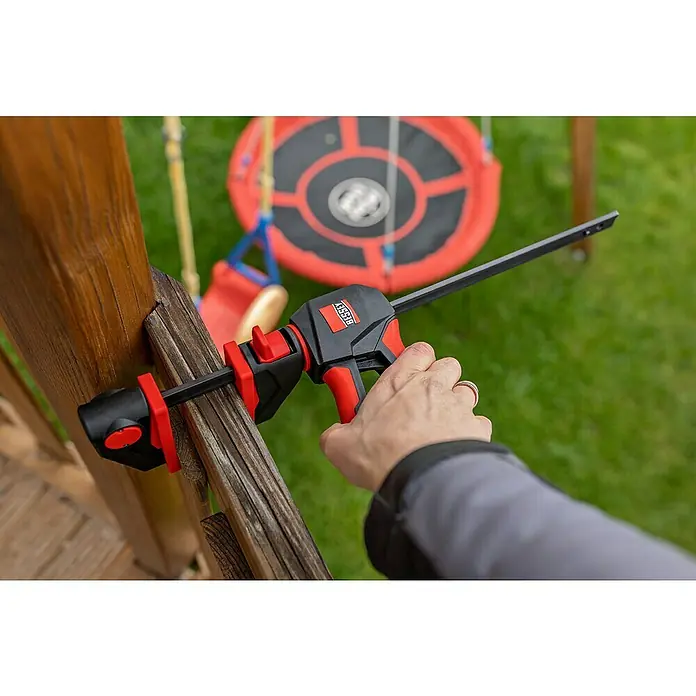 Bessey Einhandzwinge EZ 360-45 - Spannweite: 450 mm, Ausladung: 80 mm - Nahaufnahme einer BESSEY Einhandzwinge, die an einem Holzstück befestigt ist, mit einem Spielplatz im Hintergrund.